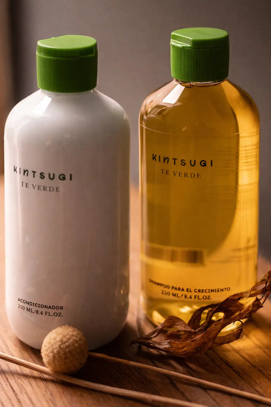 Dos botellas de kintsugi te verde, una blanca con tapa verde que contiene acondicionador y la otra botella transparente ámbar con tapa verde a juego que contiene champú para el crecimiento del cabello.
