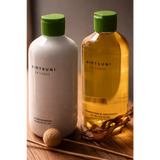 Dos botellas de kintsugi te verde, una blanca con tapa verde que contiene acondicionador y la otra botella transparente ámbar con tapa verde a juego que contiene champú para el crecimiento del cabello.